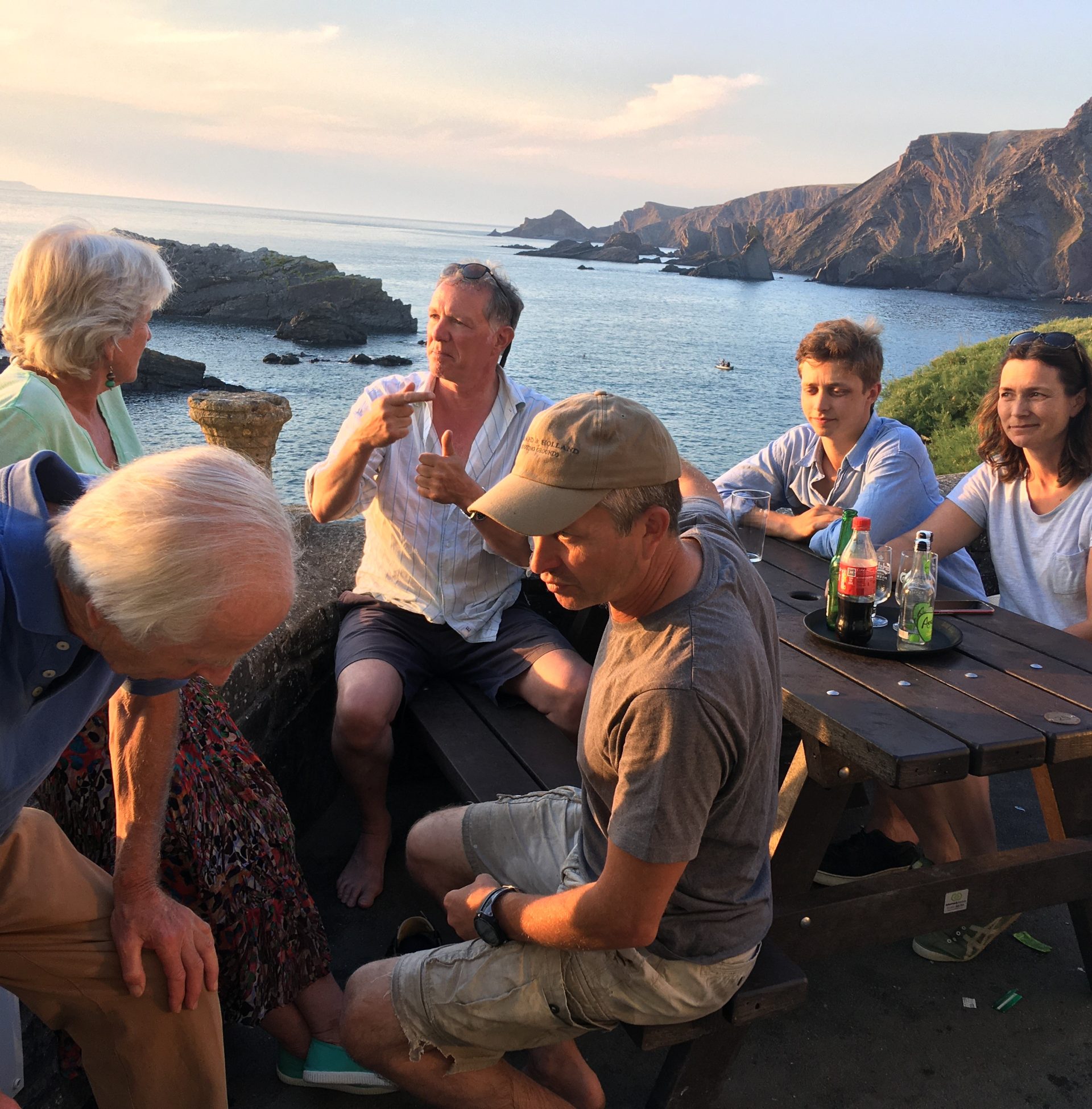Group of people gathered for food and drinks on picnic table in beer garden of Hartland Quay, with rugged coastline in background