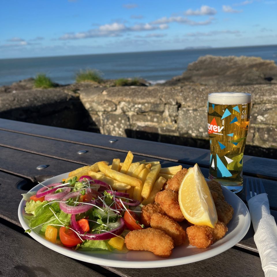 Plate of scampi and chips with pint of lager set on picnic table overlooking Hartland Quay